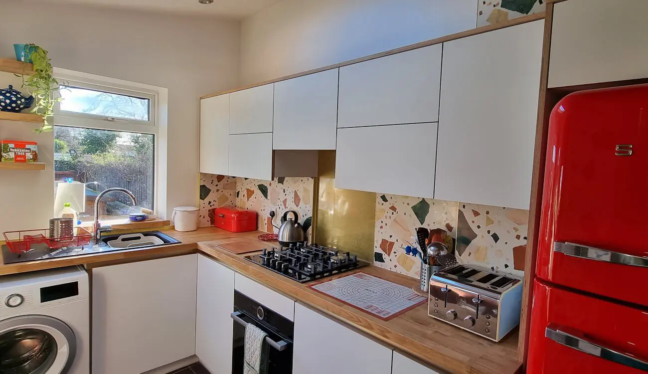White laminated birch plywood kitchen with exposed ply edges, terrazzo backsplash, and compact L-shaped layout, handcrafted by JS DECO.