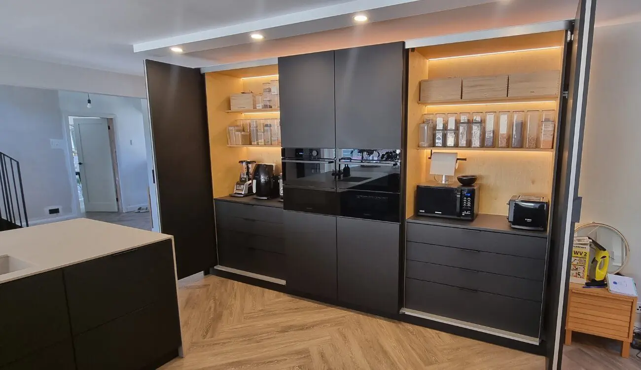 Black birch plywood kitchen with pocket-door breakfast pantry, LED-lit birch interior shelving, and integrated appliances, handcrafted by JS DECO.
