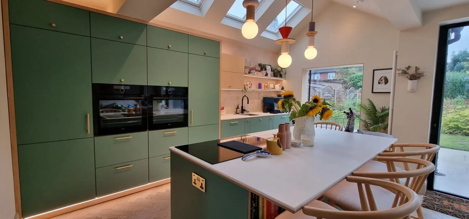 Modern bespoke plywood kitchen in matte green finish with exposed birch ply edges, featuring floor-to-ceiling custom cabinetry and a central island with seating.