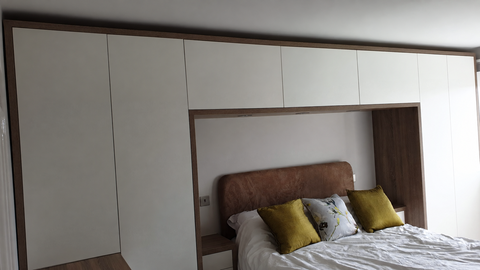 Fitted wardrobes and overhead cupboards frame the bed, with white matt fronts and an oak-effect surround.
