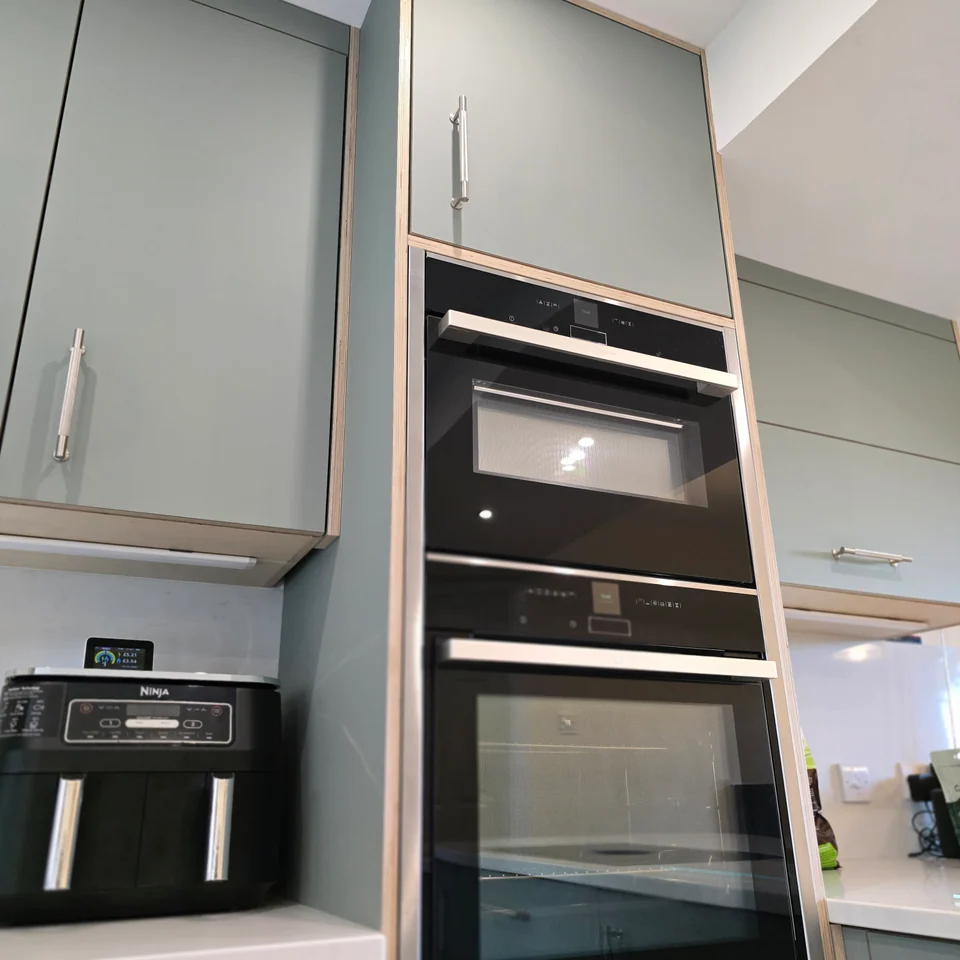 Muted green kitchen cabinetry with exposed plywood edges, metal handles and integrated ovens.