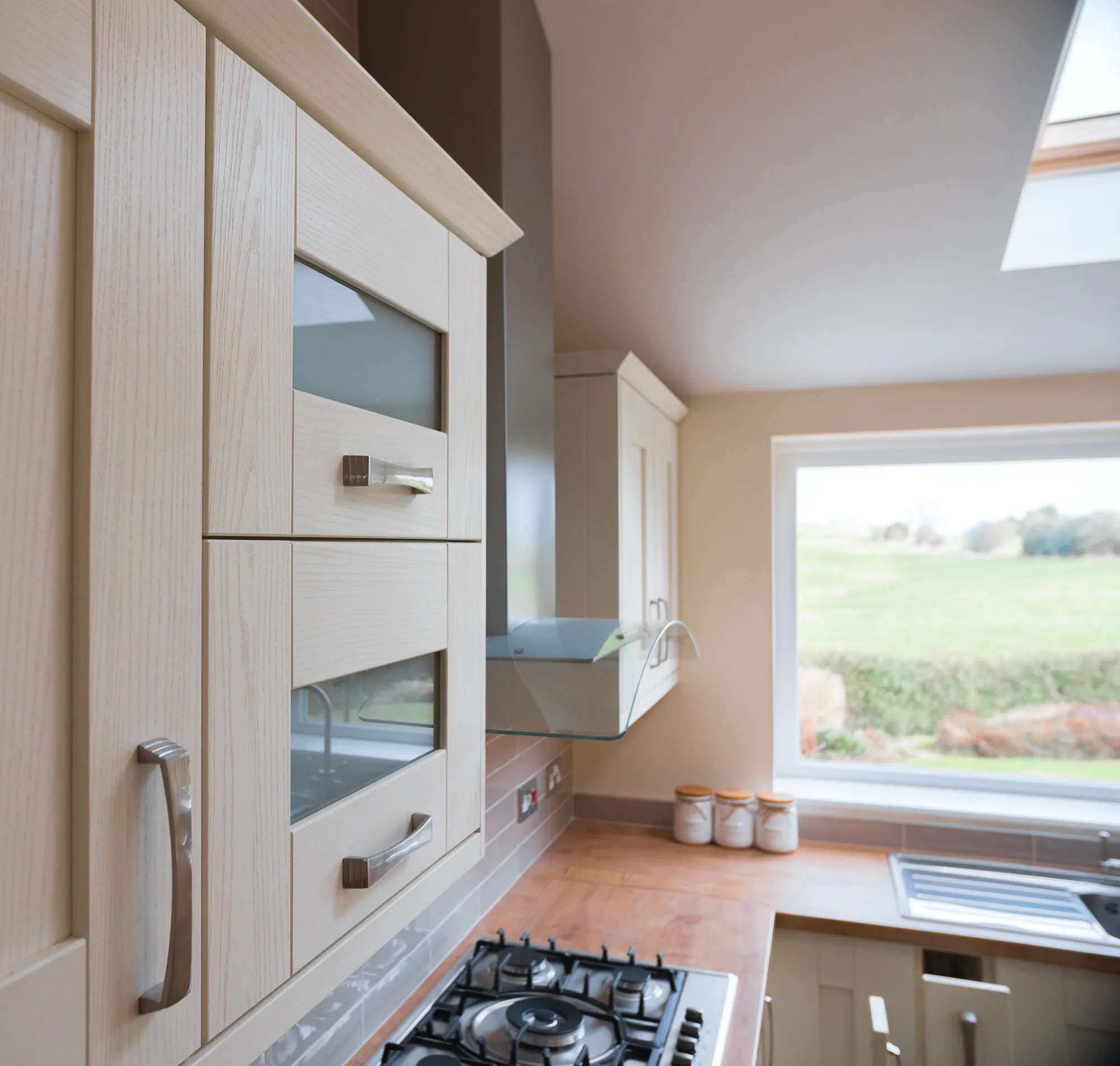 Detail of the hand-painted style Shaker wall units, hob and extractor facing the garden views. Ivory Shaker wall cabinets with stainless steel handles above gas hob and oak worktop in Pudsey Leeds kitchen.